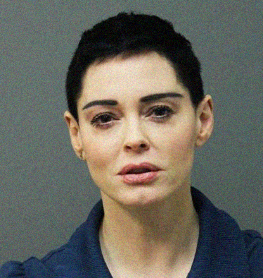 A woman with short black hair and a serious expression looks directly at the camera against a plain background. She is wearing a dark collared top.