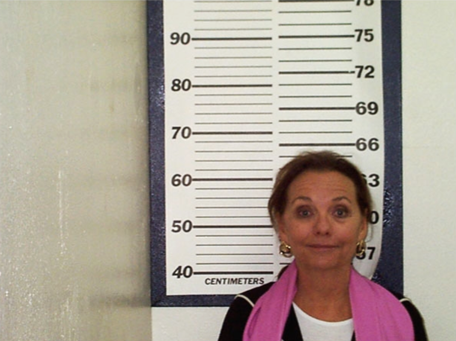 A woman stands in front of a height measurement chart marked in centimeters, smiling slightly at the camera. She wears a pink scarf and large earrings. The chart is white with black lines and numbers.