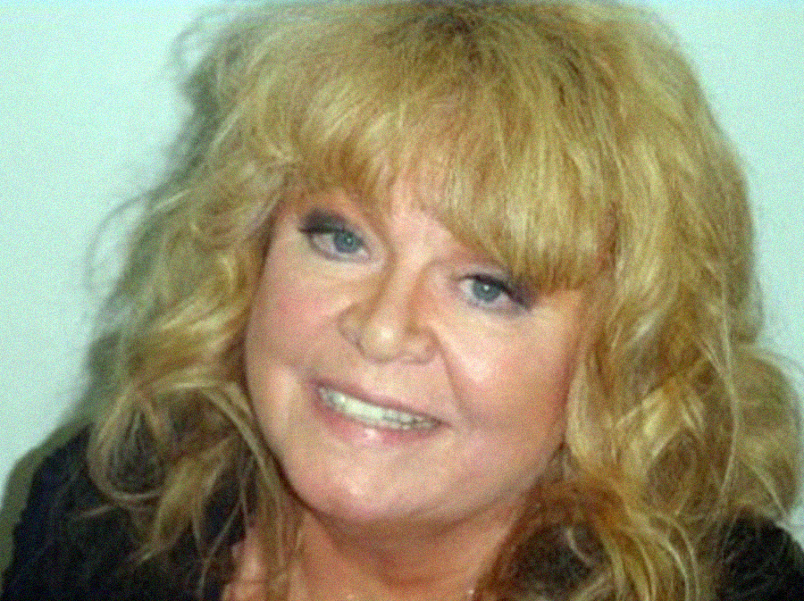 A woman with wavy blonde hair smiles at the camera. She has light skin, blue eyes, and is wearing makeup with soft pink lipstick. The background is plain and light-colored.