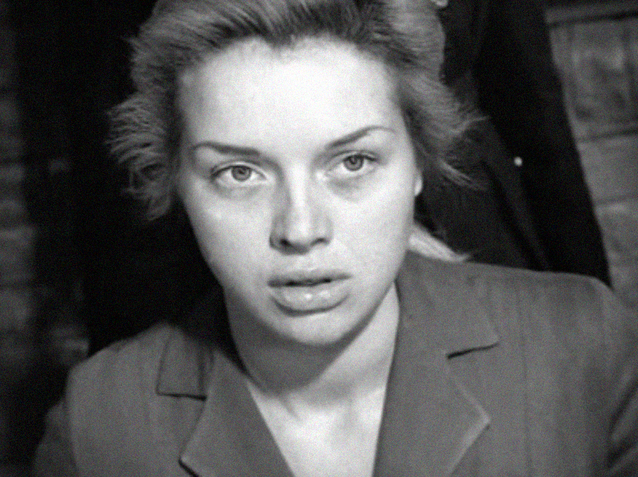 Black and white photo of a young woman with light hair, parted to the side, wearing a collared jacket. She appears to be looking slightly upward with a serious or thoughtful expression. The background is out of focus.