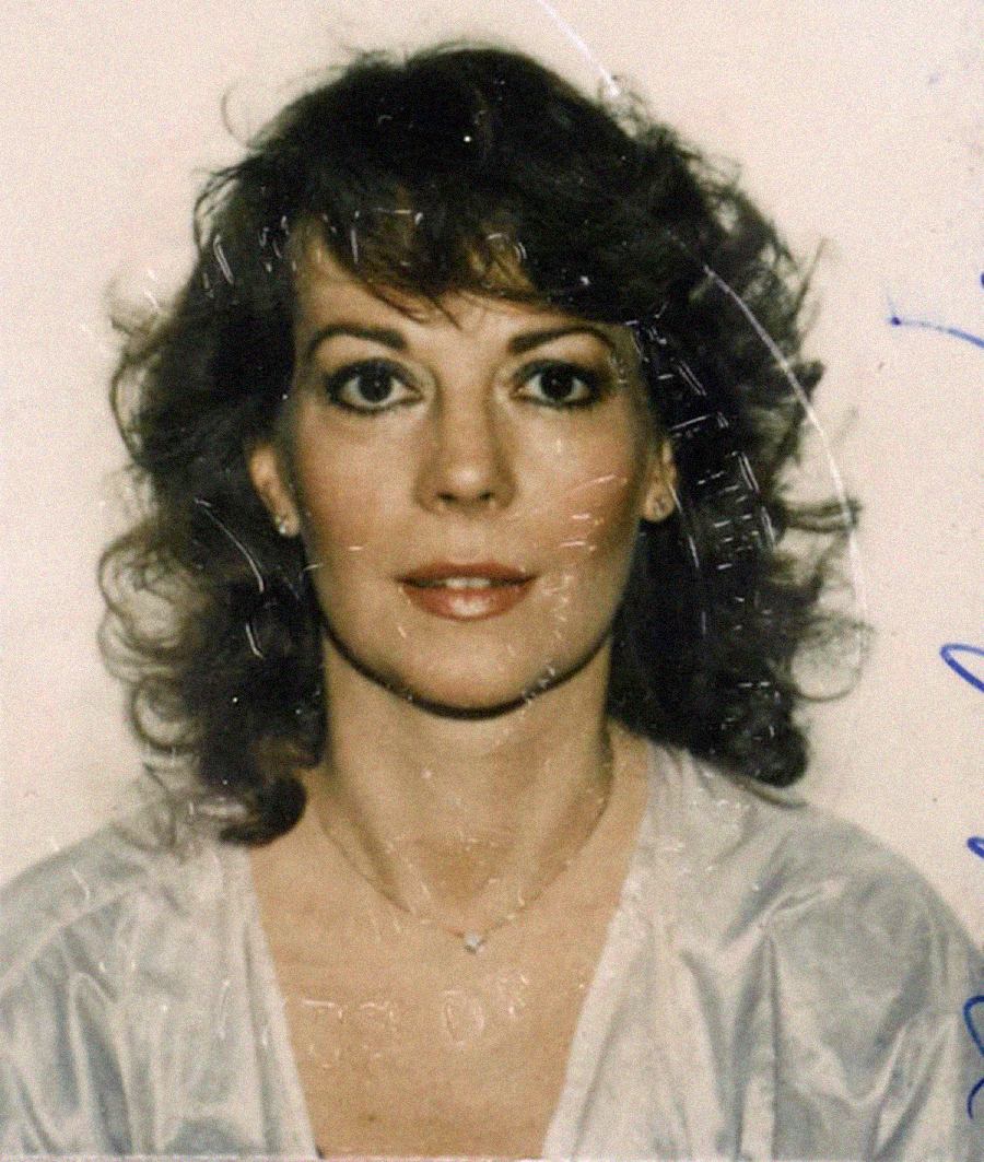 A woman with shoulder-length wavy brown hair and light makeup looks directly at the camera, wearing a light-colored top and a delicate necklace, against a plain background.