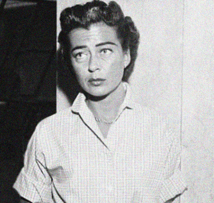 Black and white photo of a woman with short, wavy hair, wearing a light-colored collared shirt, standing against a plain background and looking slightly upward with a serious expression.