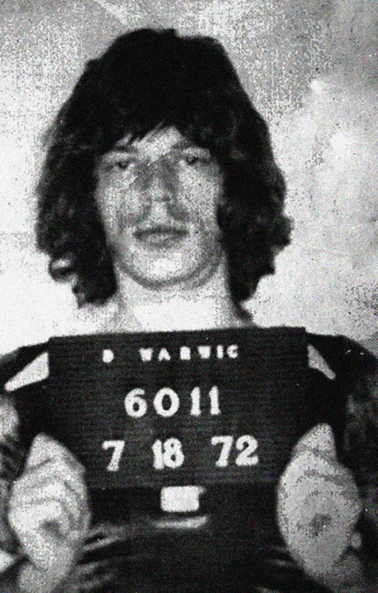 Black and white image of a person with medium-length dark hair holding a sign with numbers and the date 7-18-72, suggesting a vintage mugshot photograph.