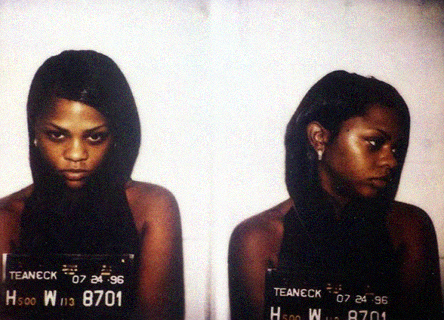 A double mugshot photo shows a woman with long dark hair, facing forward in one image and to the side in the other. She holds a placard with numbers and "Teaneck 07 24 96" written on it.