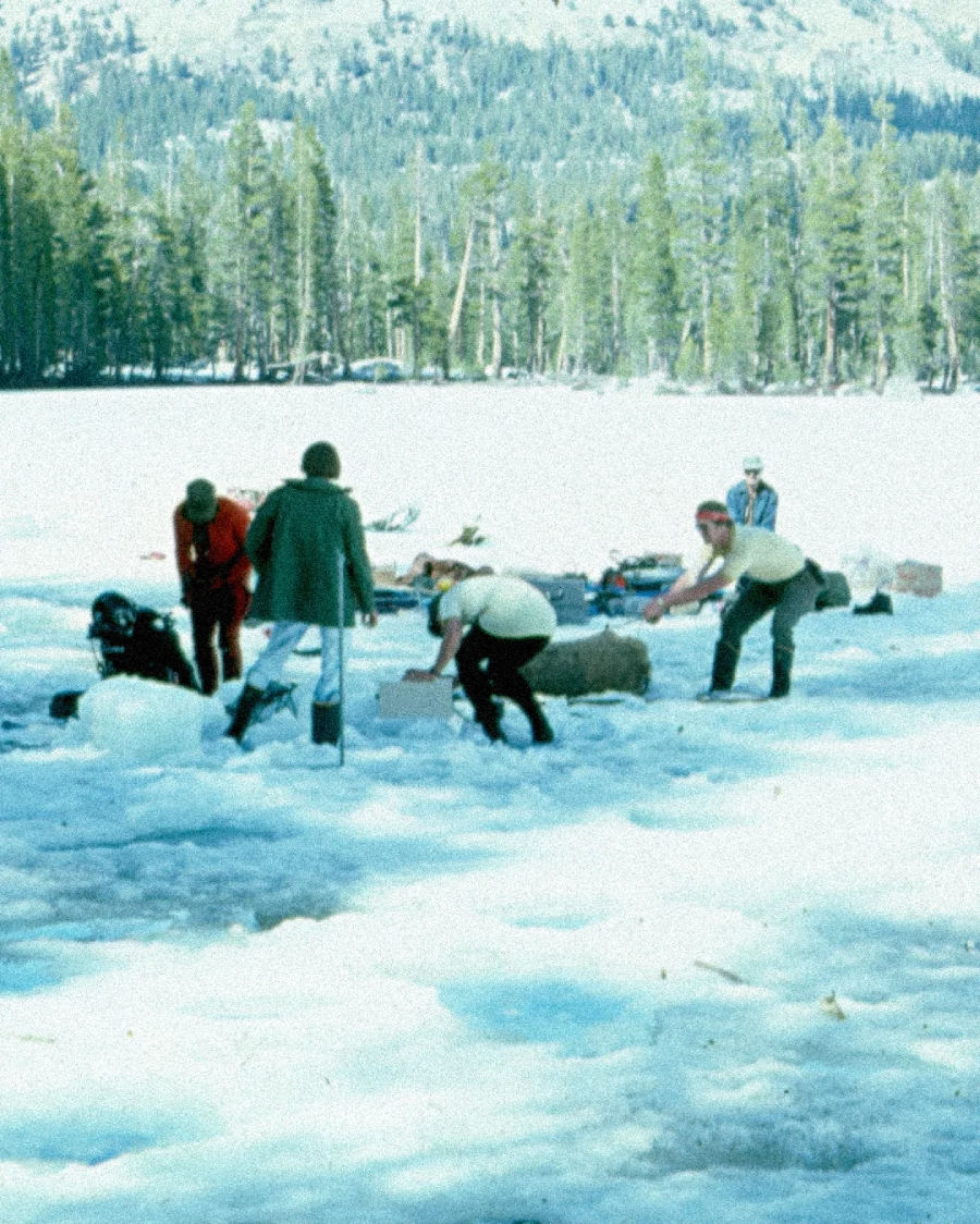 A group of people stand and work on a snowy, frozen lake surrounded by trees and distant mountains, with equipment and supplies scattered on the ice.
