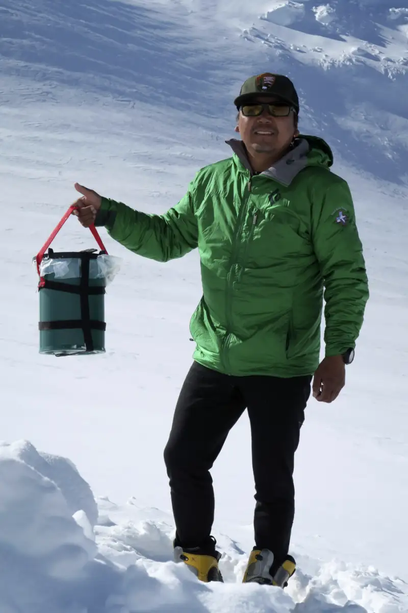 A person wearing a green jacket, black pants, boots, and sunglasses stands on snow, holding a green bucket with a red handle. Snowy, mountainous terrain is visible in the background.