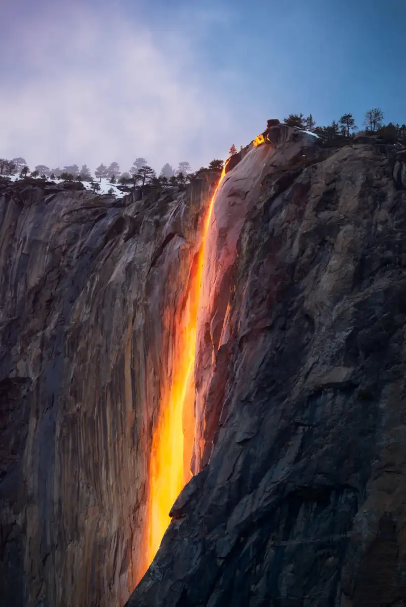 A bright orange waterfall glows like flowing lava against a rocky cliff at dusk, with trees and snow visible at the top; the sky is softly lit in the background.
