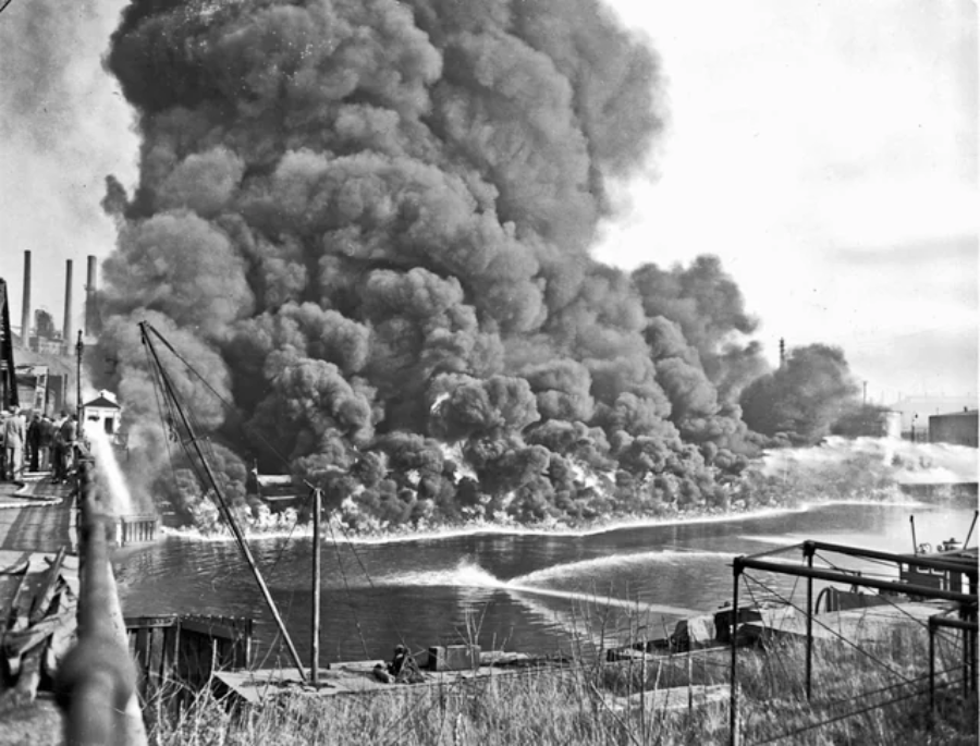 A massive cloud of dark smoke billows over a river as firefighters spray water toward the flames from the riverbank and boats. Industrial buildings and factories are visible in the background.