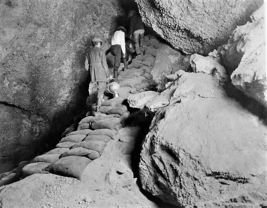 Three people climb a stairway made of sandbags inside a rocky cave or tunnel. The environment appears rough, with uneven rock walls and dim lighting.