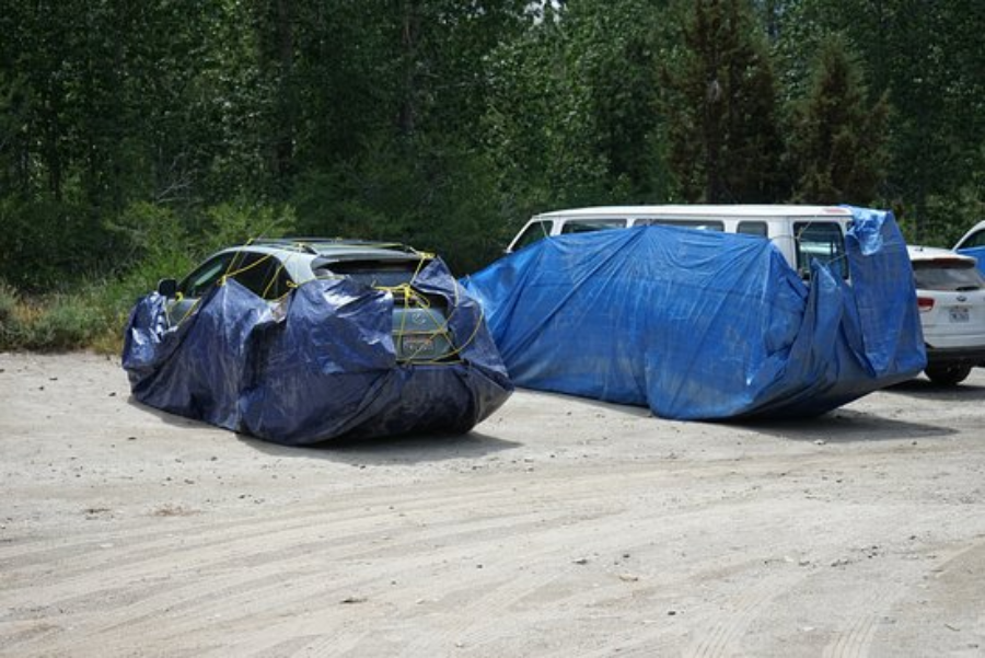 Two vehicles parked outdoors are mostly covered with blue tarps secured by yellow cords. The area is surrounded by trees, and a white SUV is partially visible in the background.