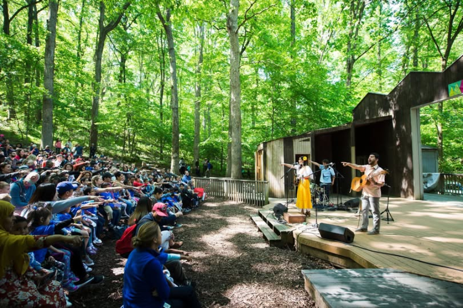 A band performs on an outdoor stage surrounded by trees while a diverse audience, including children, sits on benches, watching and interacting with the musicians during the day.