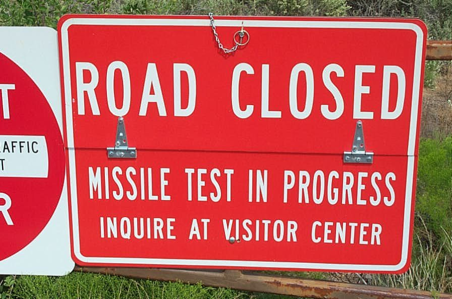 A red sign reads "Road Closed. Missile Test In Progress. Inquire at Visitor Center," warning drivers about an active missile test and road closure. Green plants are visible in the background.