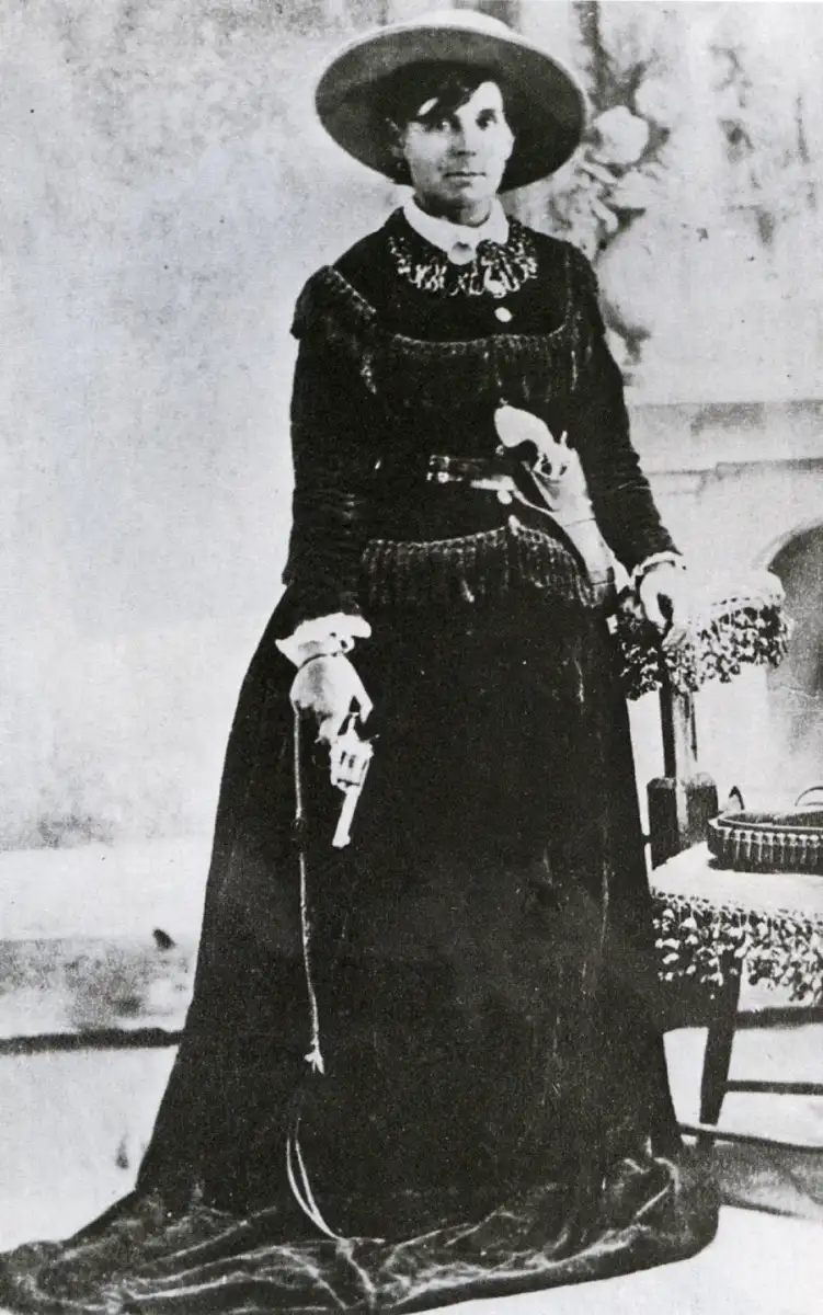 A woman in a long dark dress, wide-brimmed hat, and necklace stands beside a chair, holding a revolver in her right hand. The photo is black and white and appears to be from the late 19th or early 20th century.