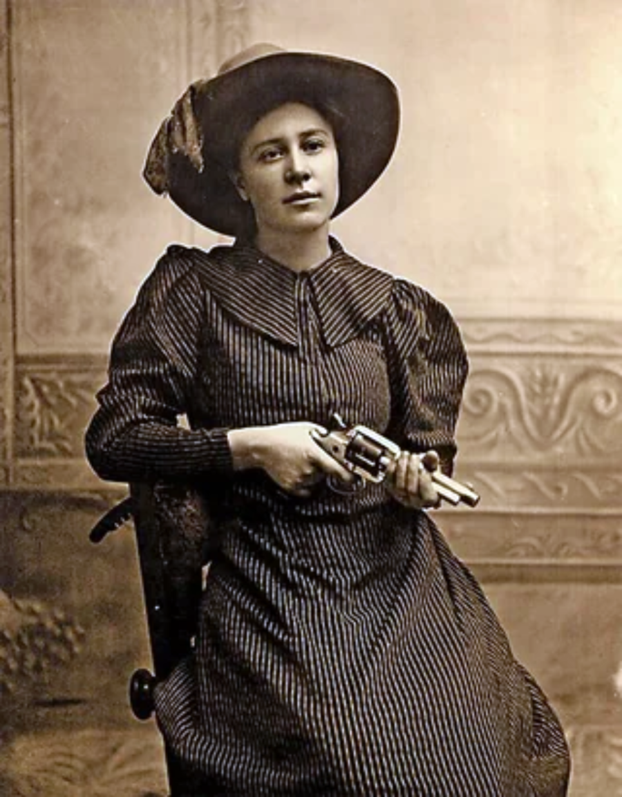 A woman in a striped dress and wide-brimmed hat sits on a chair, holding a revolver across her lap. The photo is sepia-toned and has a vintage, old West appearance.