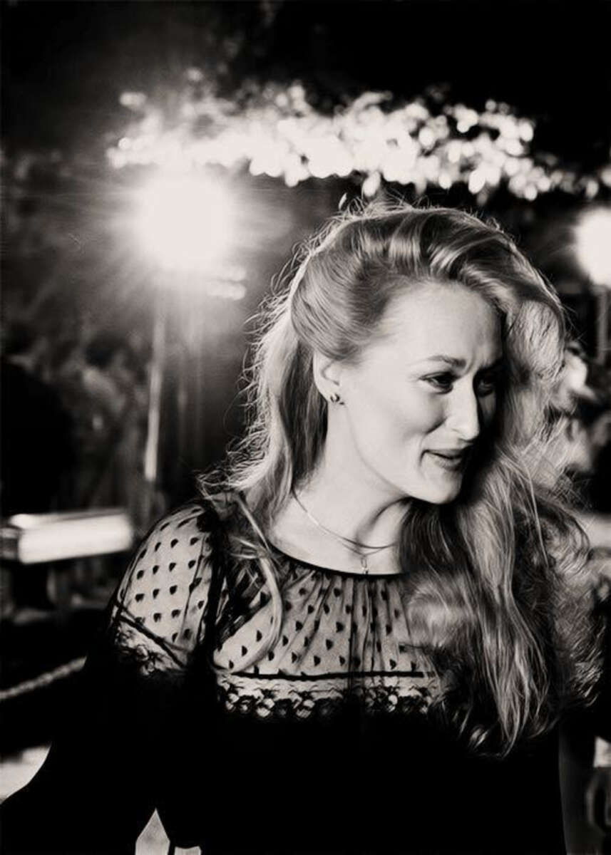 A woman with long, wavy hair and a sheer, polka-dot top smiles as she stands outdoors at night, with bright lights and blurry trees in the background.
