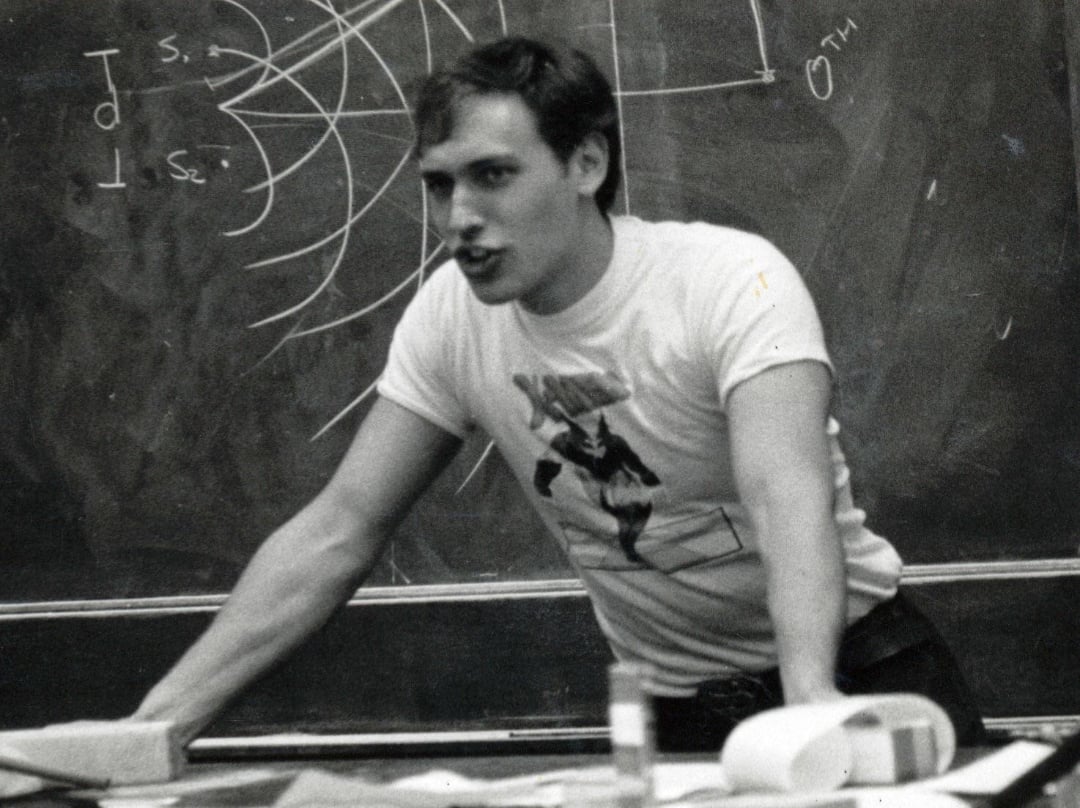 A young man in a graphic t-shirt leans forward with both hands on a desk, speaking passionately in front of a chalkboard covered with diagrams and equations.