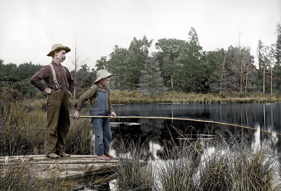 Two boys stand barefoot on a log by a pond, holding fishing poles. They wear old-fashioned clothes and hats, surrounded by tall grass and trees, gazing intently at the water.