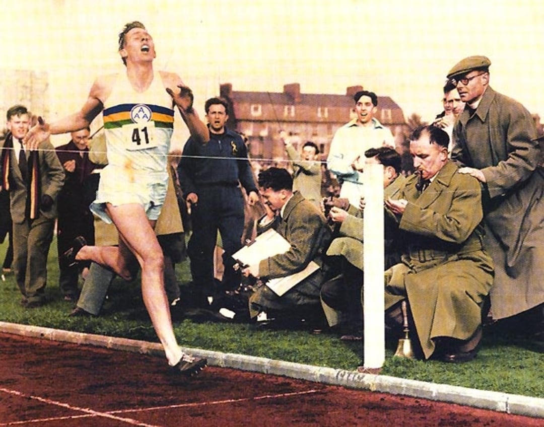 A runner wearing number 41 crosses the finish line on a track, with spectators and photographers capturing the moment. The scene appears to be from a historic athletic event, with men in coats watching excitedly.