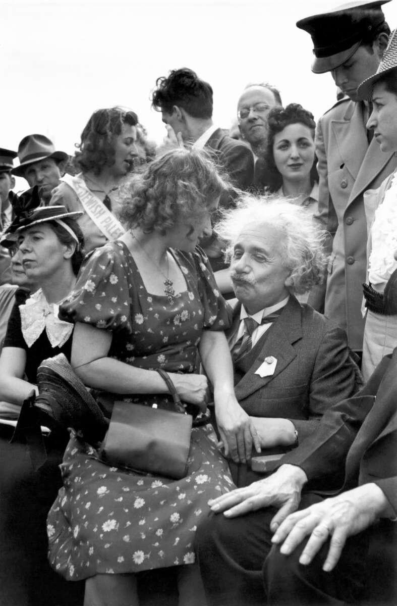 A man with wild white hair, wearing a suit, sits among a crowd. A woman in a floral dress sits on his lap, and people around them are smiling and talking, creating a lively, cheerful scene.