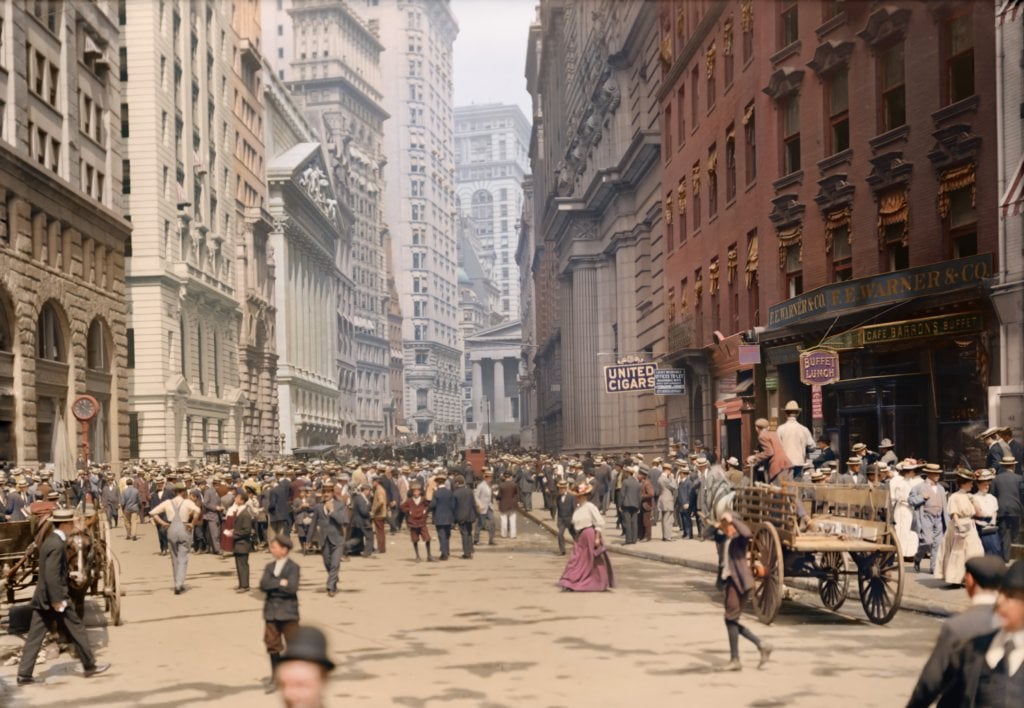 A busy city street scene from the early 1900s shows crowds of people in period clothing, horse-drawn carts, and tall buildings lining both sides of the street. Signs for shops and businesses are visible.