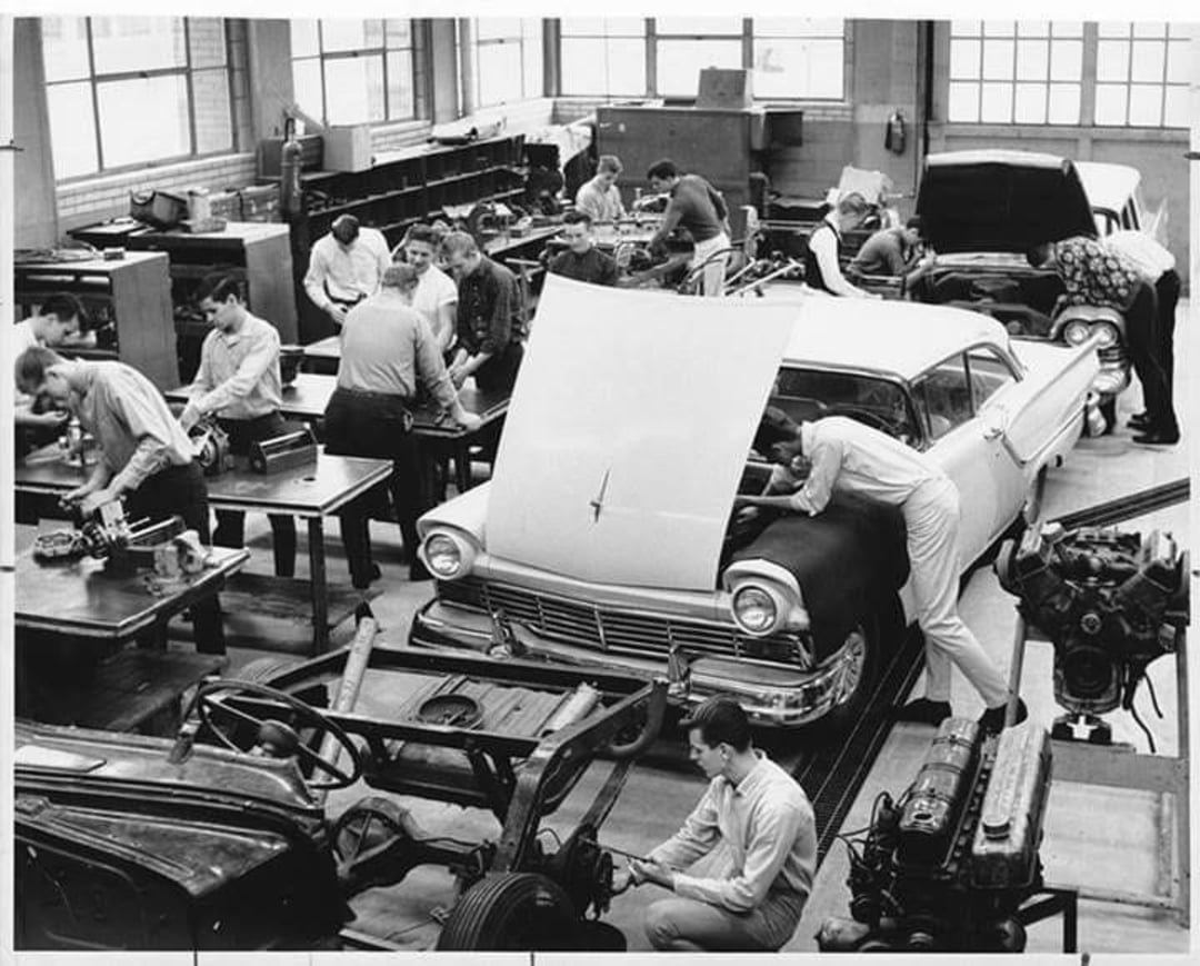 Black-and-white photo of several people, mostly men, working on car engines and automotive parts in a busy workshop or classroom, with a vintage car in the center and tools scattered around.