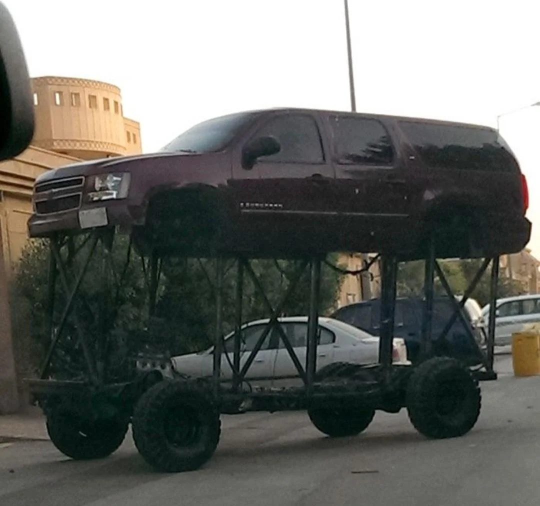 A maroon SUV is mounted unusually high on a custom, elevated chassis with large wheels, towering above a regular car parked nearby on a city street.