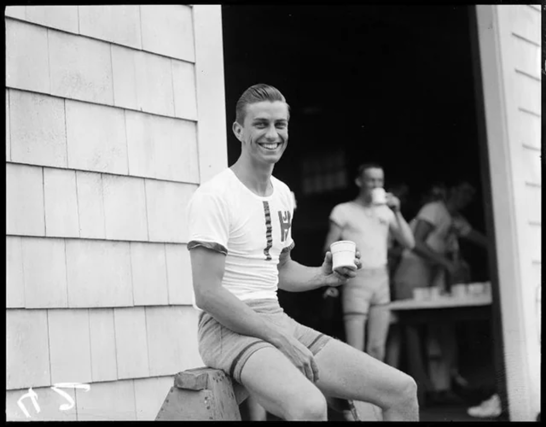 A young man in athletic clothing sits and smiles at the camera while holding a cup. Other people in similar attire are gathered inside a building behind him, some also holding cups. It appears to be a casual, outdoor setting.