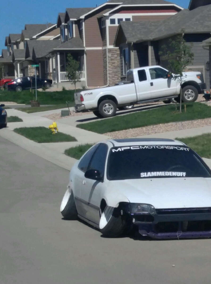 A white car with extreme negative camber and lowered suspension is parked on a residential street. Nearby, houses and a white pickup truck are visible. The car's windshield displays "MPC MOTORSPORT" and "SLAMMEDENUFF" decals.