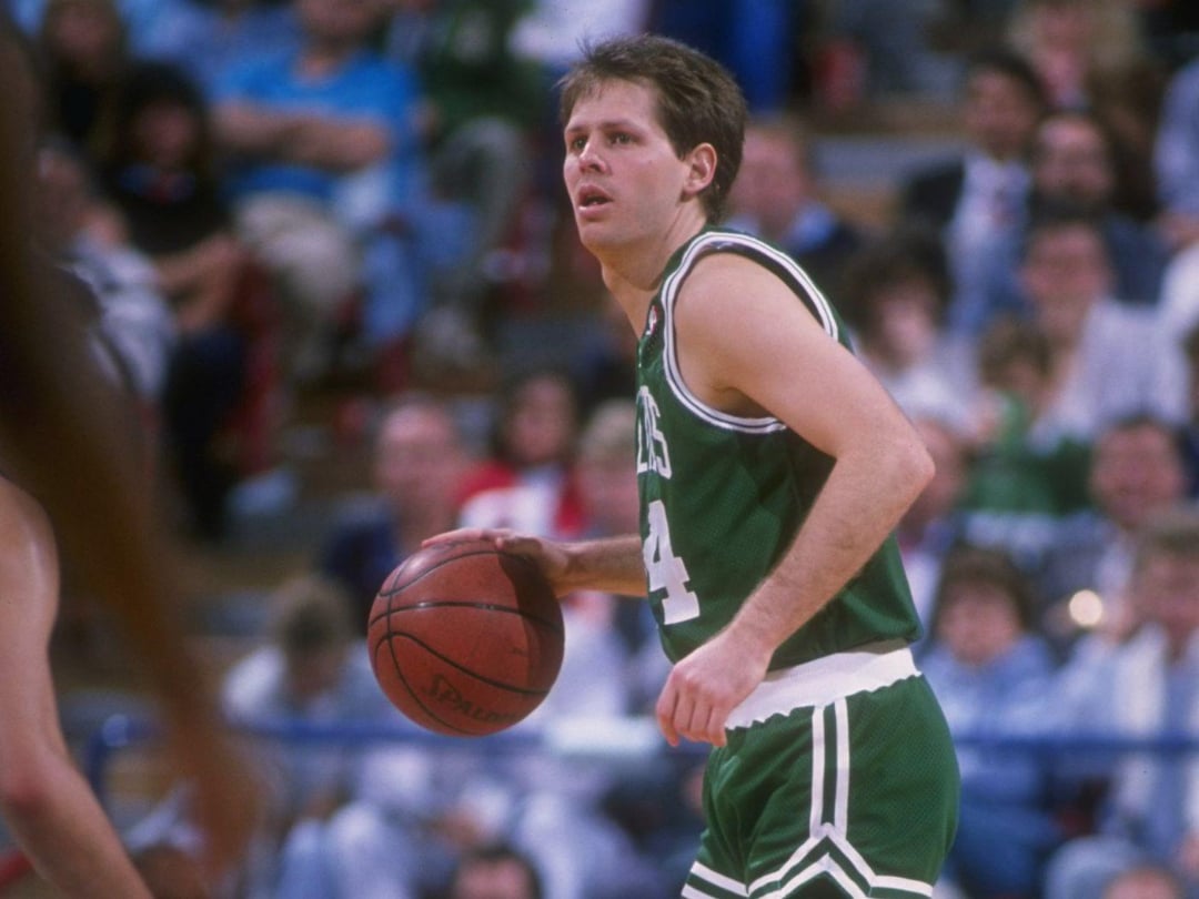 A basketball player in a green and white uniform dribbles the ball on a court, with an audience blurred in the background.
