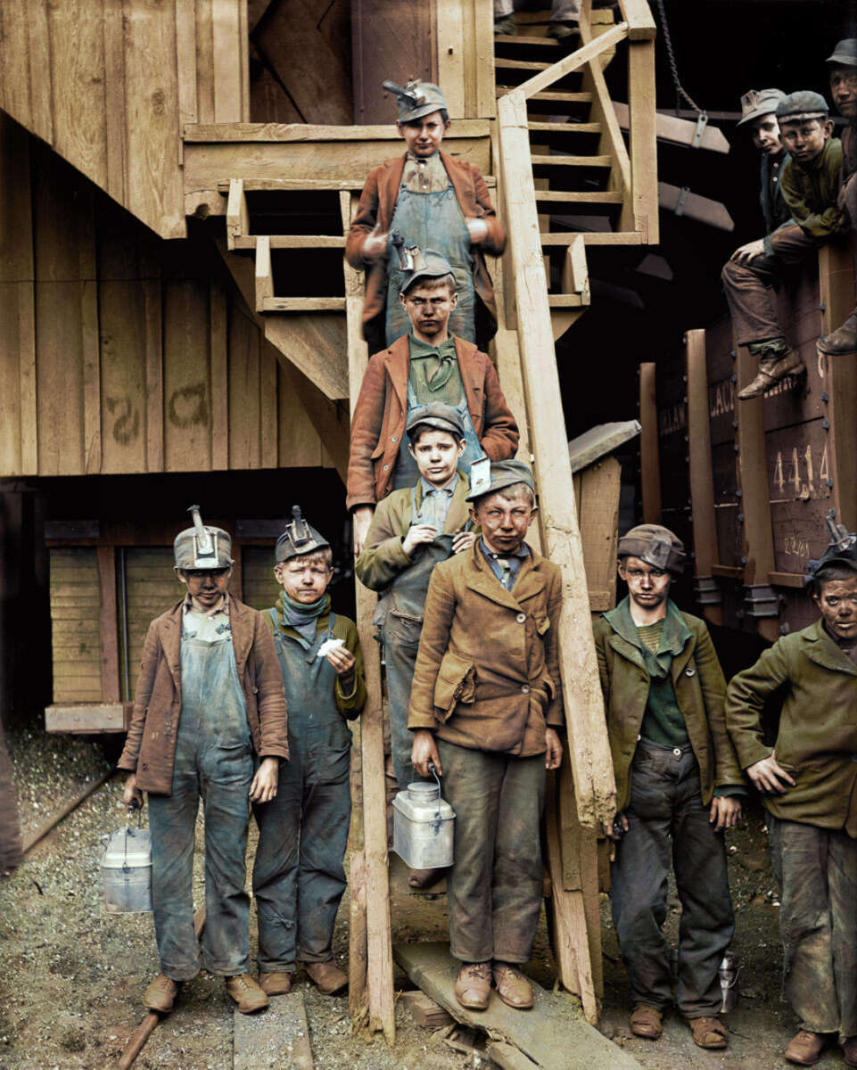 A group of young boys wearing work clothes and headlamps stand and sit on wooden stairs outside a coal mine, holding metal lunch pails. Their faces are dirty, and the background shows wooden structures and a railcar.