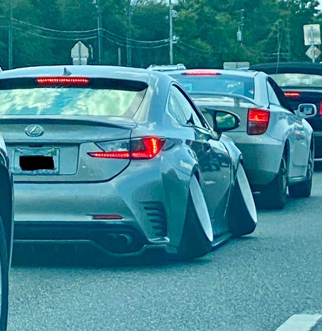 A silver Lexus car with extremely tilted wheels, known as “stance” or “cambered” style, waits in traffic behind other vehicles on a road. The car's tires angle sharply inward at the bottom.