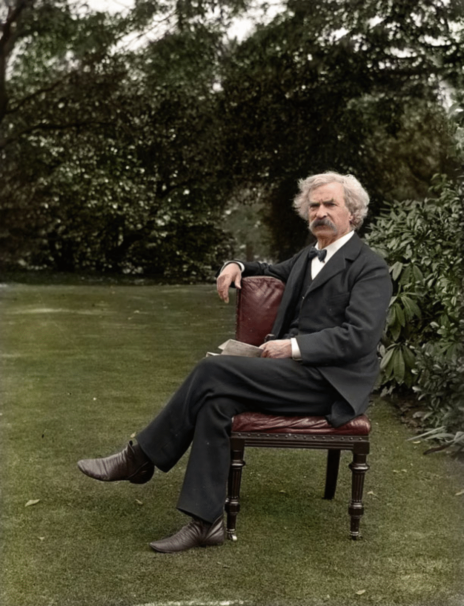 An older man with white hair and a mustache sits on a wooden chair in a garden, wearing a dark suit and holding a book. Trees and bushes fill the background.