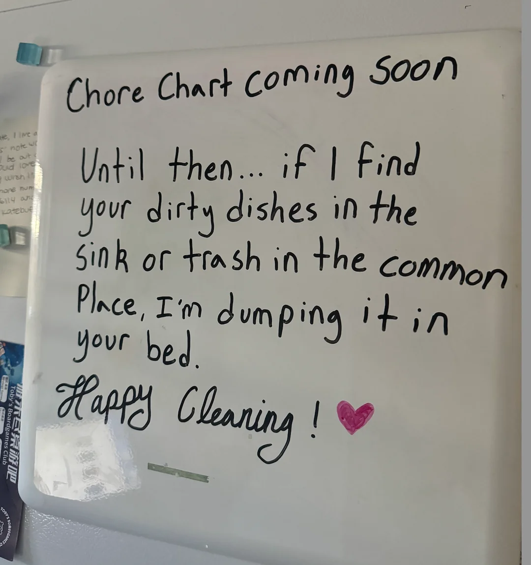 A whiteboard on a fridge reads: "Chore chart coming soon. Until then... if I find your dirty dishes in the sink or trash in the common place, I'm dumping it in your bed. Happy cleaning!" with a pink heart drawn.