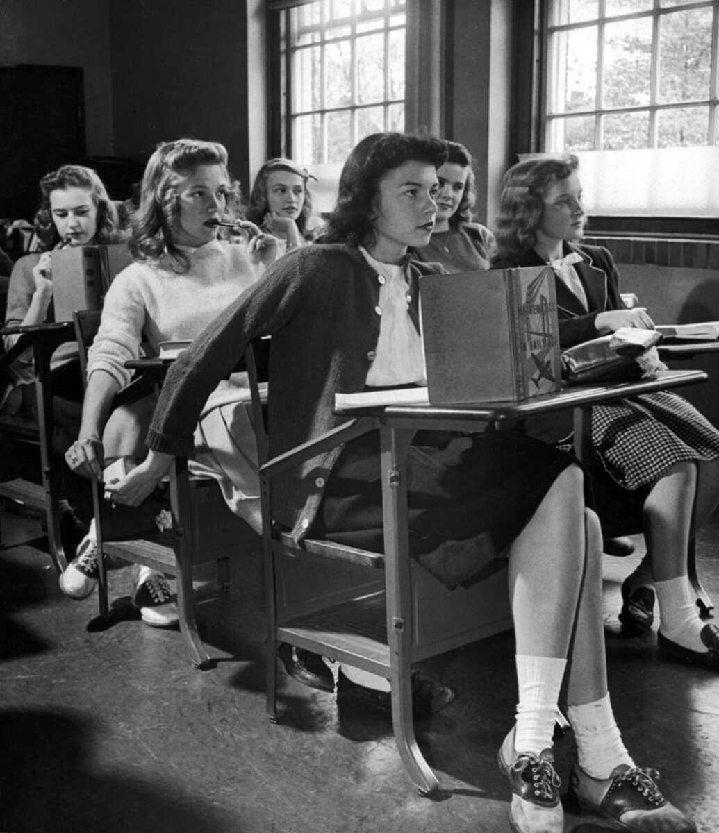 Six teenage girls sit in a classroom at old-fashioned desks, some passing notes behind their backs, while others look forward or sideways. Sunlight streams through large windows, creating a bright, vintage atmosphere.