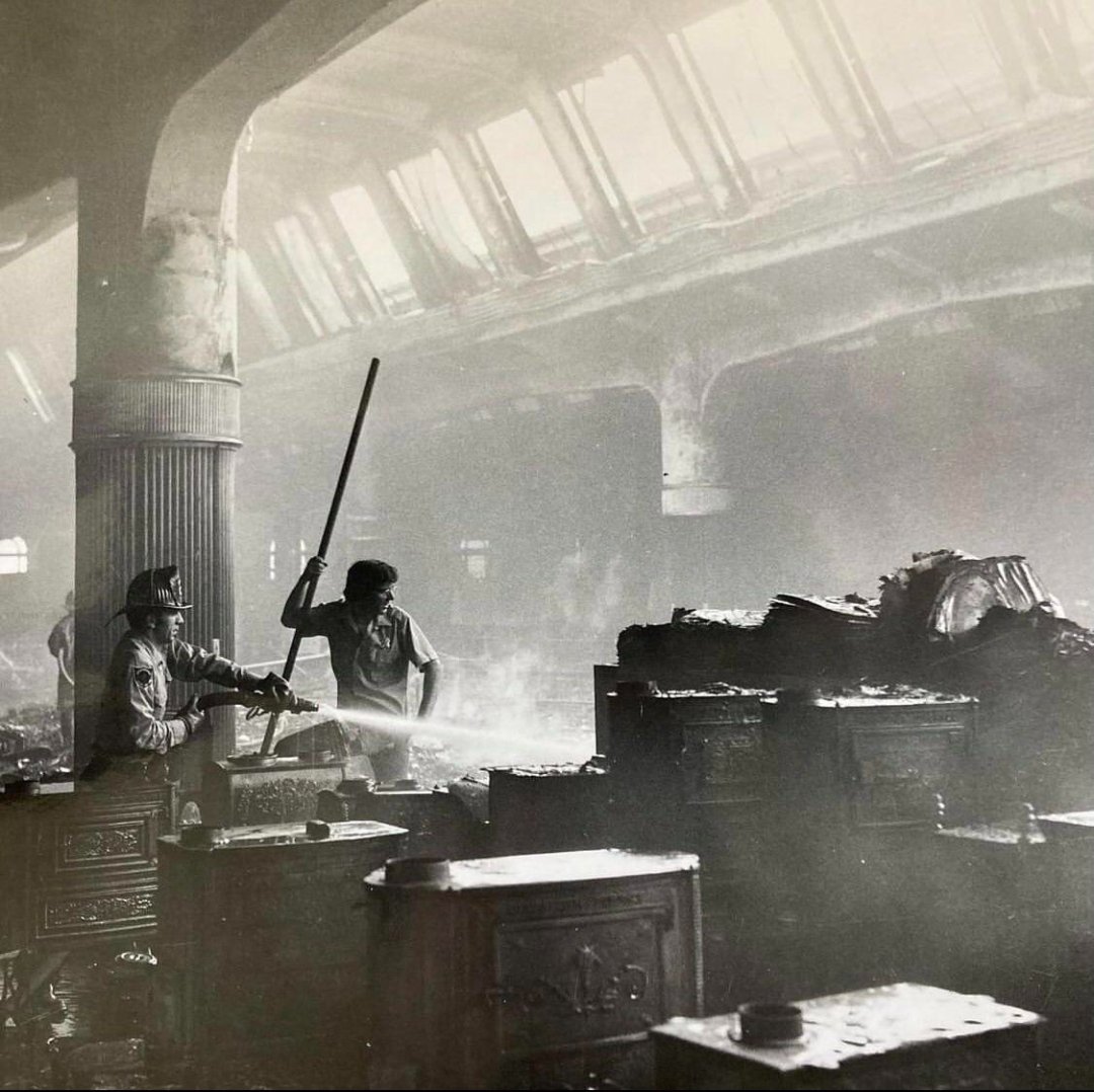 Two firefighters spray water inside a smoke-filled, partially destroyed building with charred debris and furniture, illuminated by light streaming through large windows above.