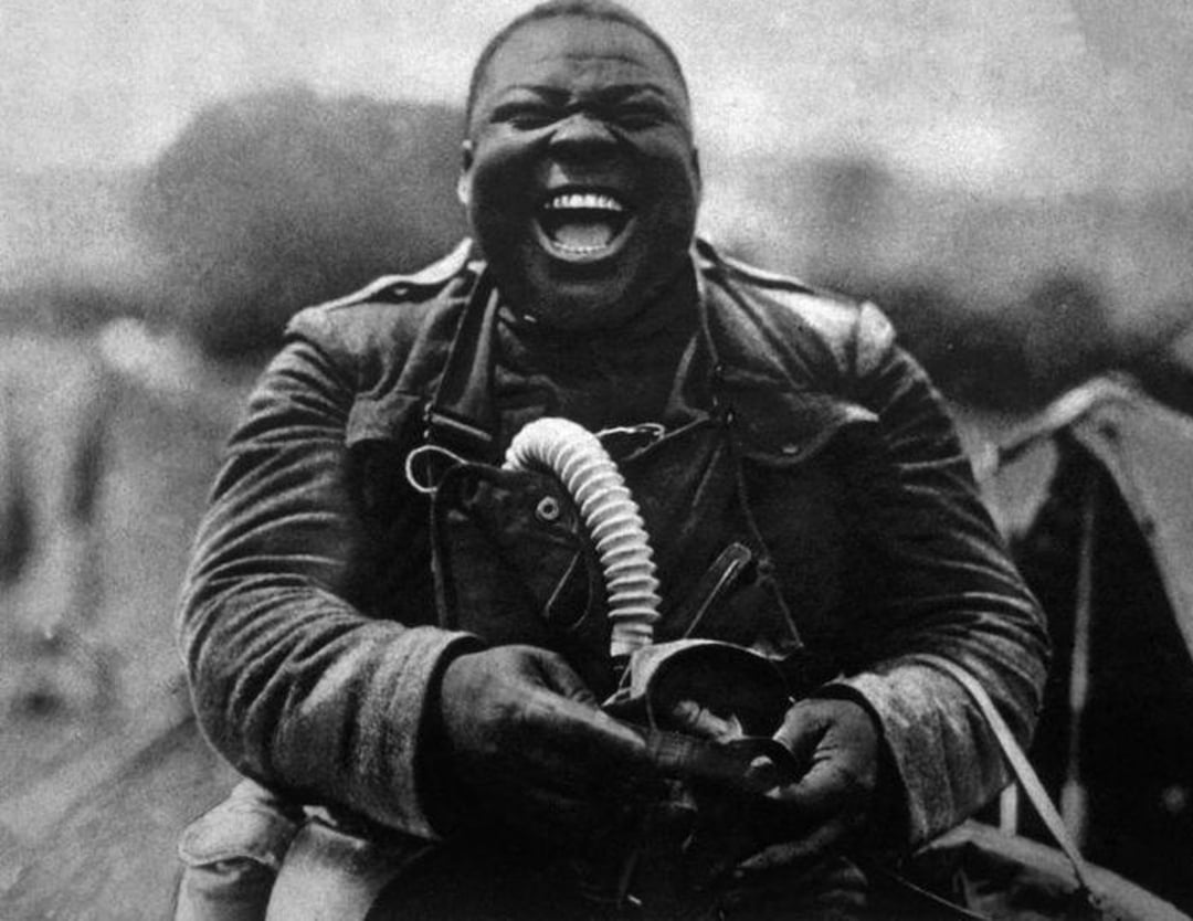 A Black World War I soldier in uniform laughs heartily while holding a gas mask, seated outdoors with a blurred background of equipment and tents.