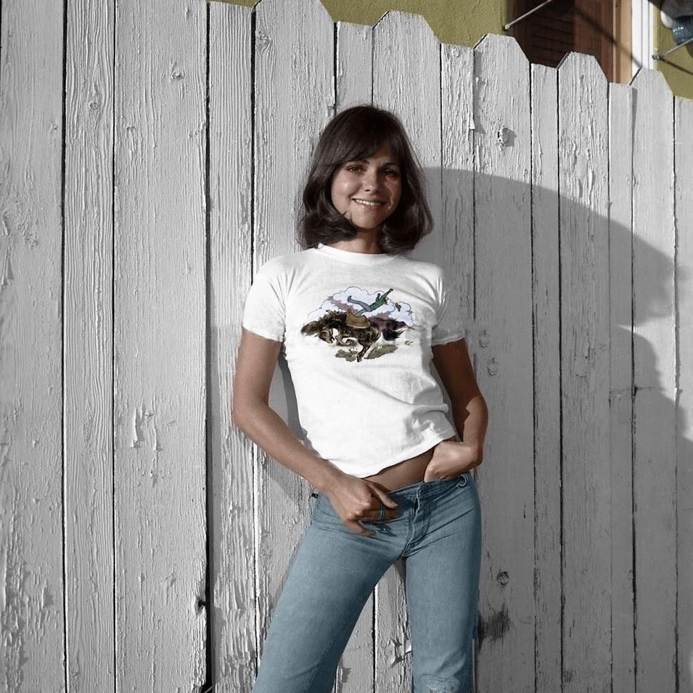 A young woman with shoulder-length brown hair stands in front of a white wooden fence. She is smiling, wearing a white t-shirt with a graphic and blue jeans, with one hand in her pocket.