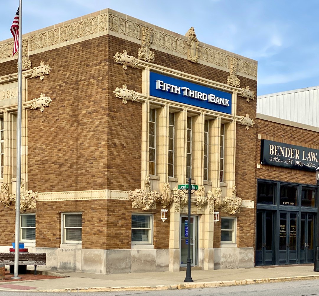 A historic brick building on a street corner houses a Fifth Third Bank branch. Decorative stonework frames the windows, and a nearby business sign reads "Bender Law." An American flag stands outside.