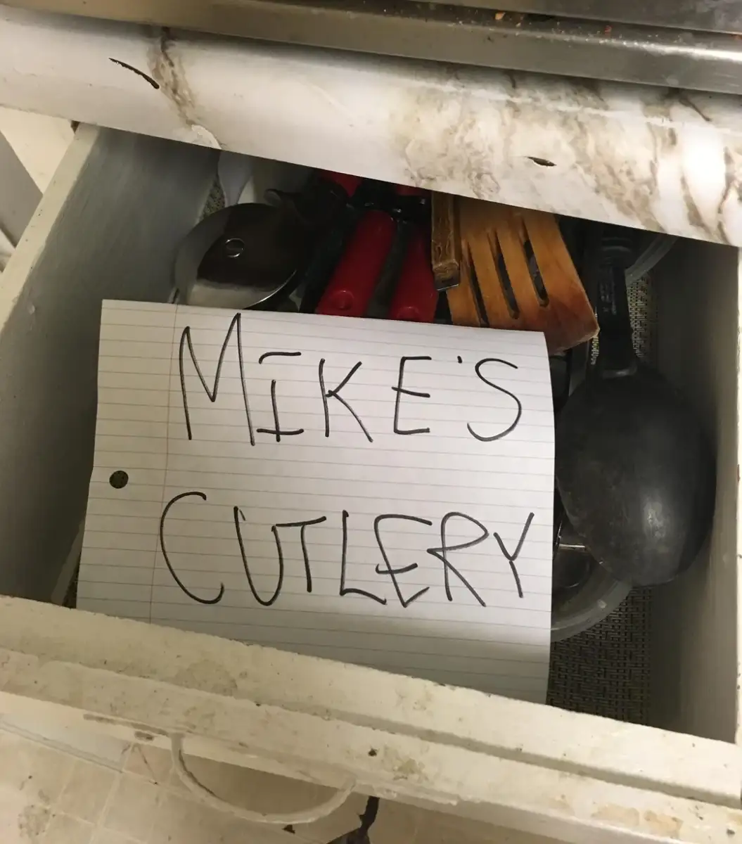 An open kitchen drawer filled with various utensils and a handwritten note on lined paper that reads "MIKE'S CUTLERY" in large letters. The drawer and surrounding area appear worn and stained.