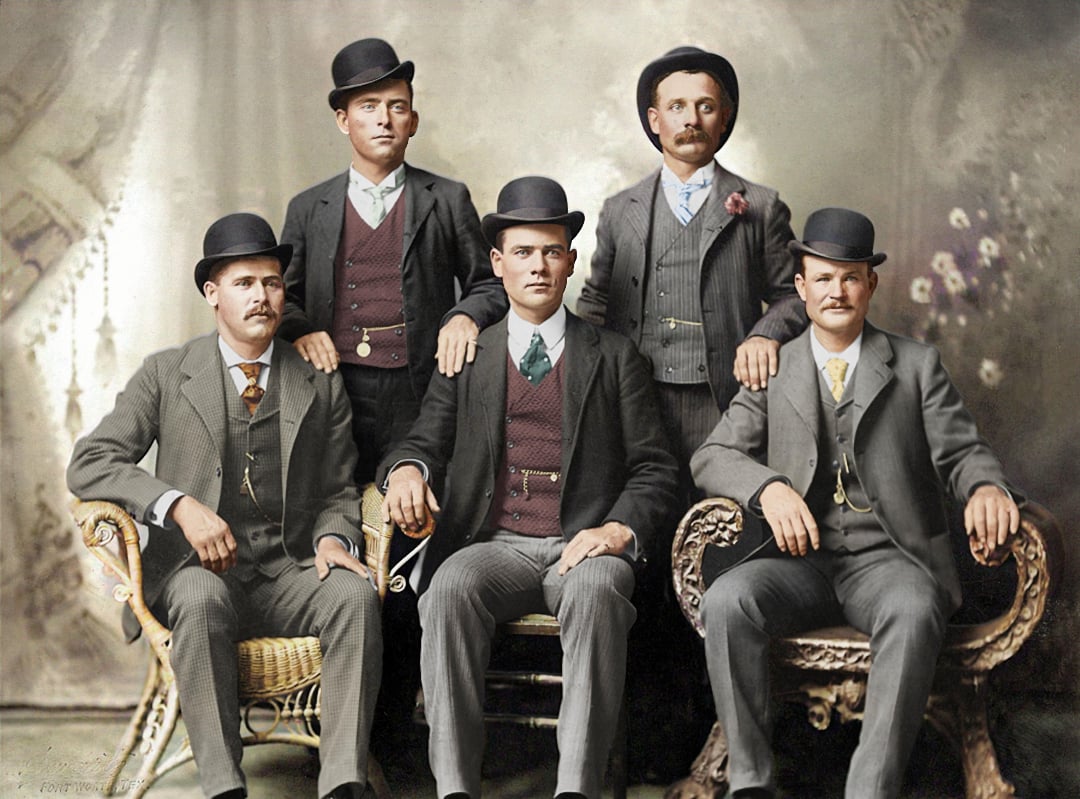 Five men in early 1900s attire, wearing suits and bowler hats, sit and stand in a formal studio portrait with a vintage painted backdrop.