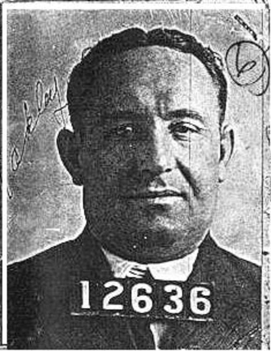Black and white mugshot of a man wearing a suit, with the number 12636 displayed on a plaque. There is a handwritten signature and the number 6 in a circle on the right side of the photo.