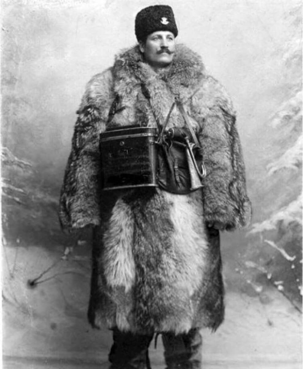 A man stands in front of a studio backdrop wearing a thick fur coat, fur hat, and a mustache. He has a drum strapped across his chest. The image is in black and white and appears old-fashioned or historical.