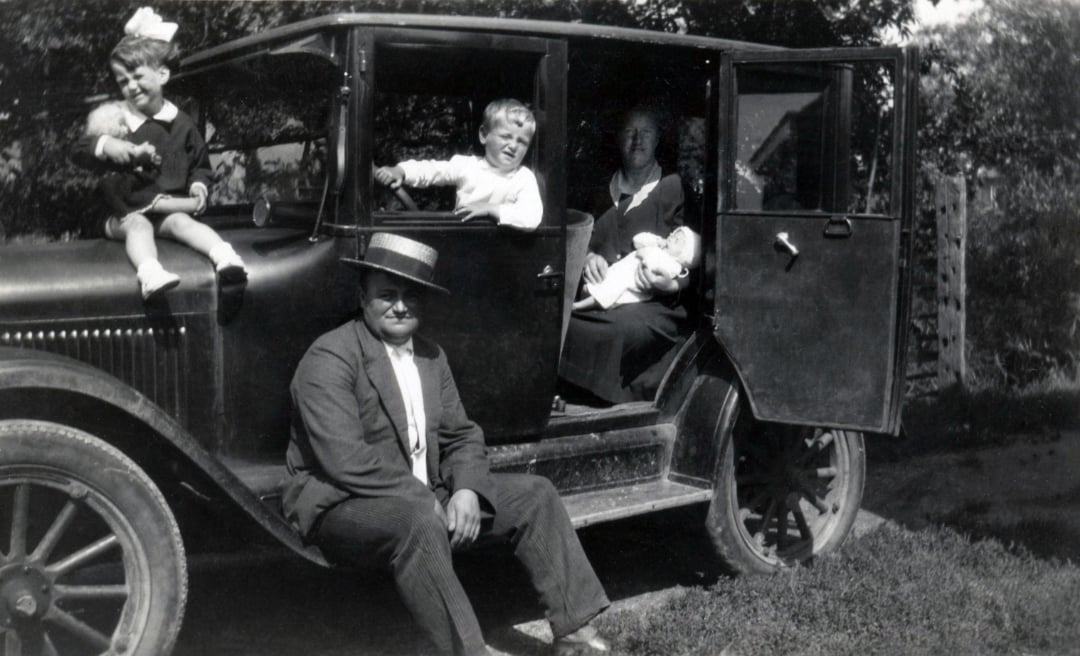 A black-and-white photo of a man in a suit and straw hat sitting on a running board of an old car. A woman, baby, and two young children are in and around the car, all posing and smiling outdoors.
