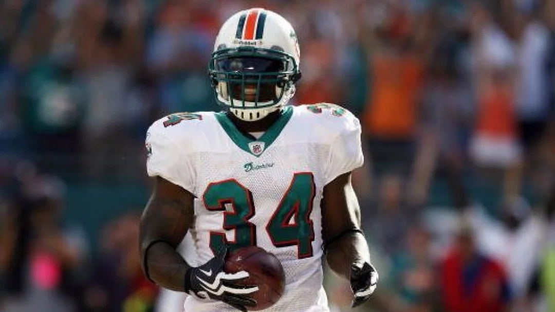 A football player wearing a Miami Dolphins uniform with the number 34 holds a football and stands on the field, with fans cheering in the blurred background.