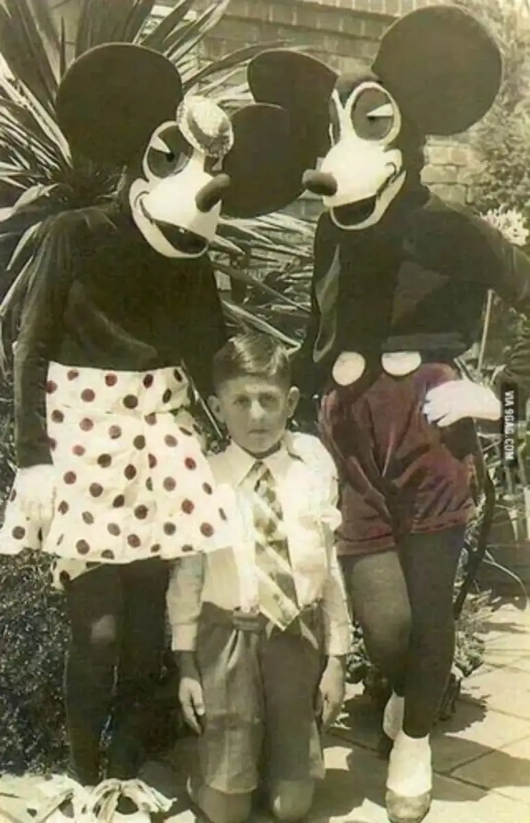 A vintage black-and-white photo shows a boy kneeling between two people in oversized handmade mouse costumes with large heads and exaggerated facial features, standing outdoors near plants.