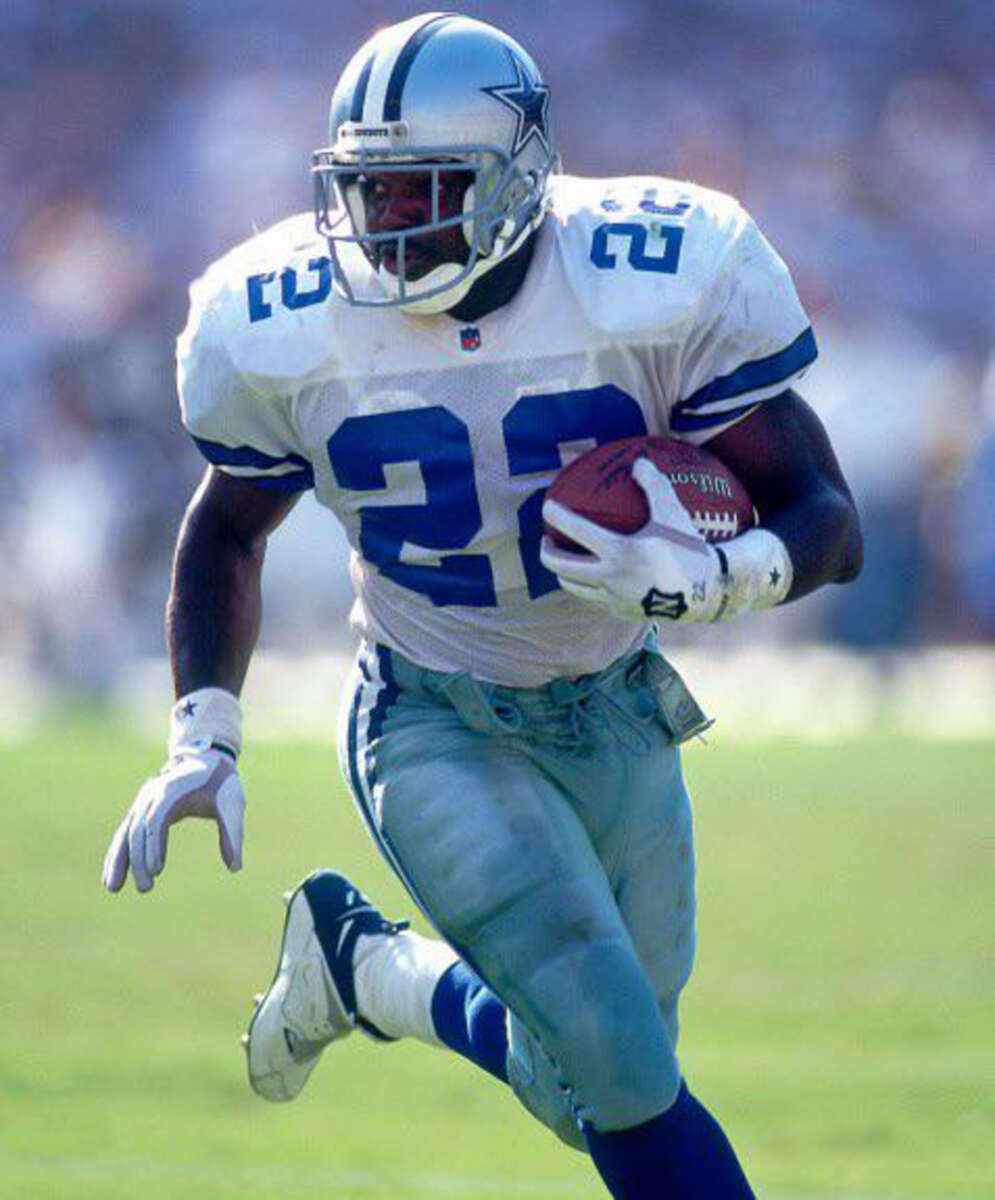 A football player in a Dallas Cowboys uniform with the number 22 runs with the ball on a sunny field, focused and mid-stride during a game.