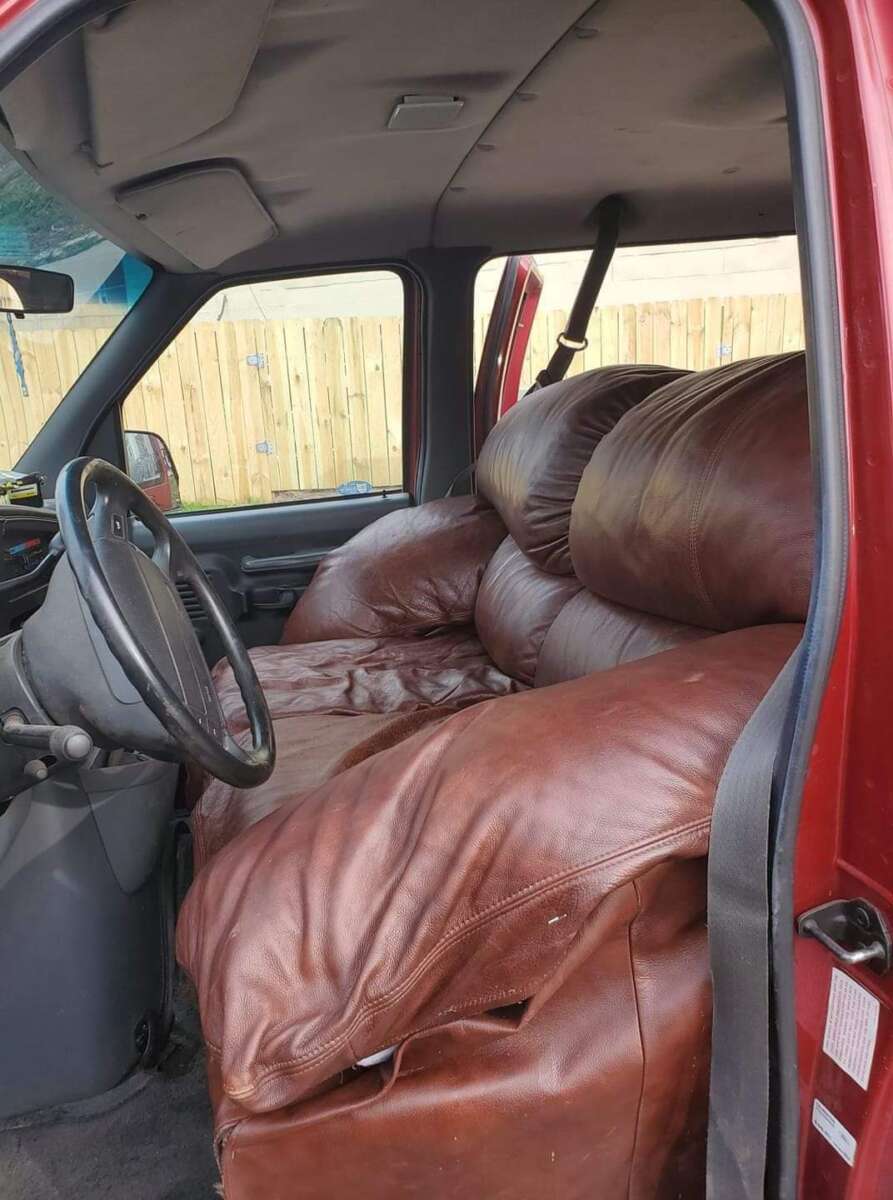 The interior of a red vehicle with a large brown leather couch replacing the front seats, creating an unusual and bulky seating arrangement. The car door is open, revealing the setup.