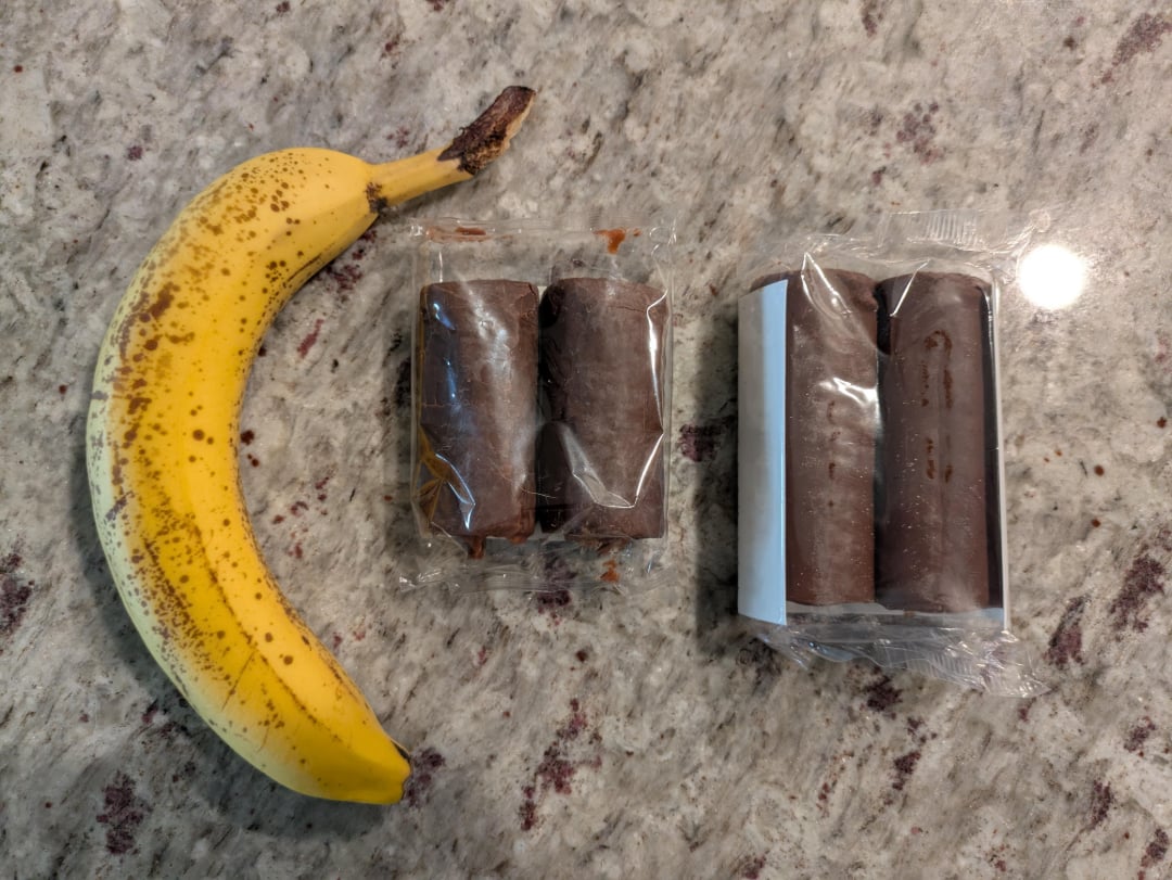 A ripe banana lies on a granite countertop next to two packs of chocolate-covered snacks, each containing two cylindrical treats wrapped in clear plastic.