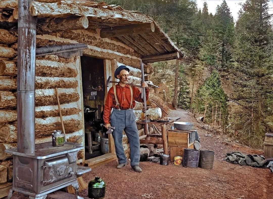 A man in suspenders and a beret stands outside a rustic log cabin holding an axe over his shoulder, surrounded by tools, firewood, and a metal stove, with a forested mountain slope in the background.
