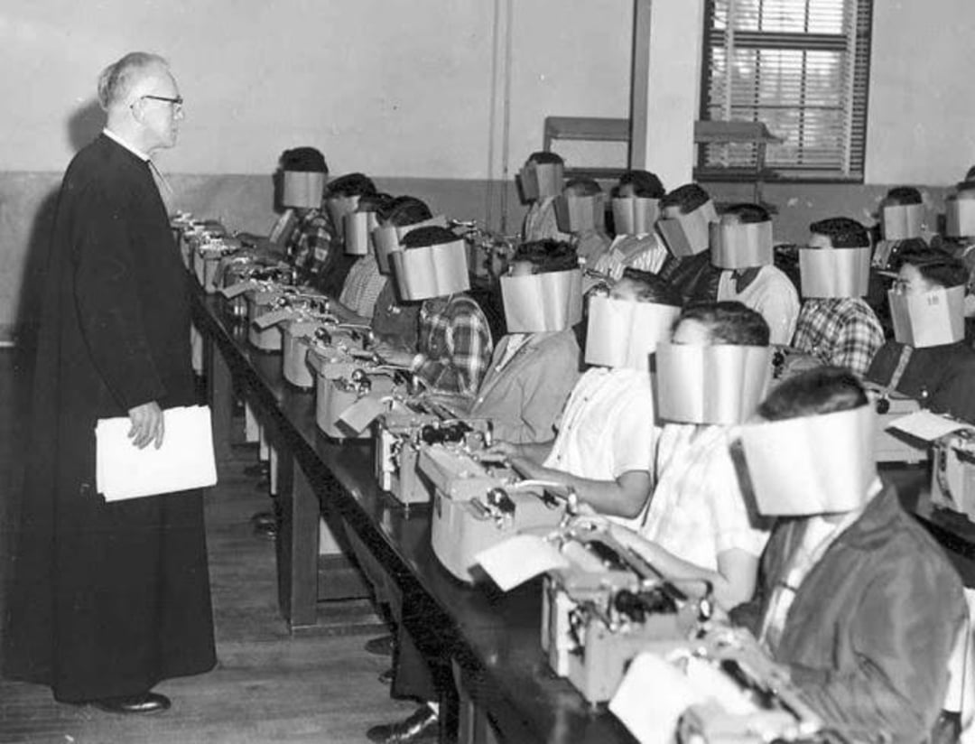 A classroom with students wearing large paper blinders on their heads, sitting at desks with typewriters, while a teacher stands at the front holding papers.